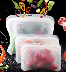 Three clear reusable silicone food bags in different sizes filled with fresh vegetables and cherry tomatoes.
