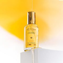 d'Alba Vita serum with vitamin capsules on a sleek surface, golden liquid dripping from the bottle against a vibrant yellow background.